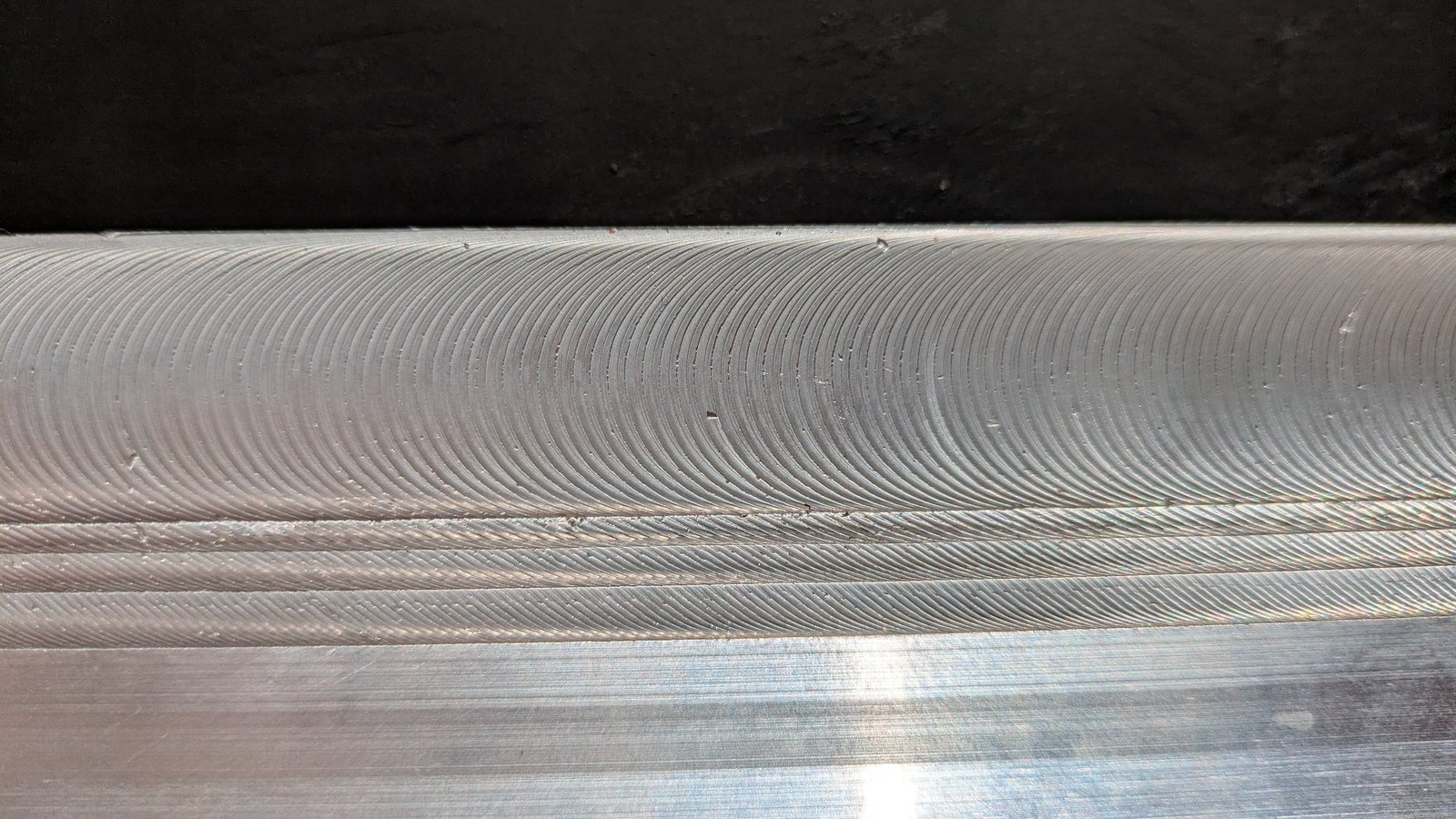 Close-up of an aluminum plate with four stacked passes of B-FSD material along its edge, with curved tool rotation marks across the top pass