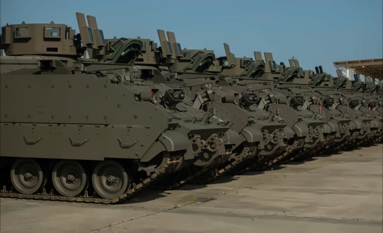 Row of olive-drab armored personnel carriers parked side by side on a concrete lot