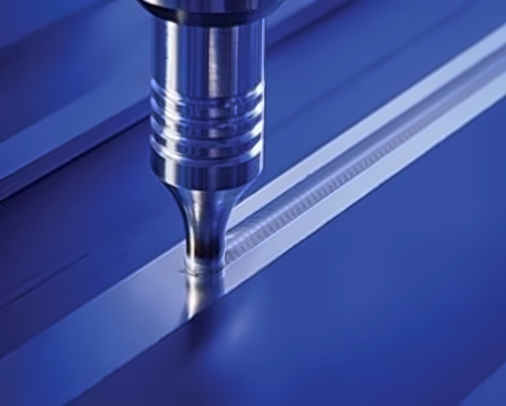 Close-up of a friction stir welding tool traveling along a seam in aluminum plate, leaving a fine ripple pattern behind it
