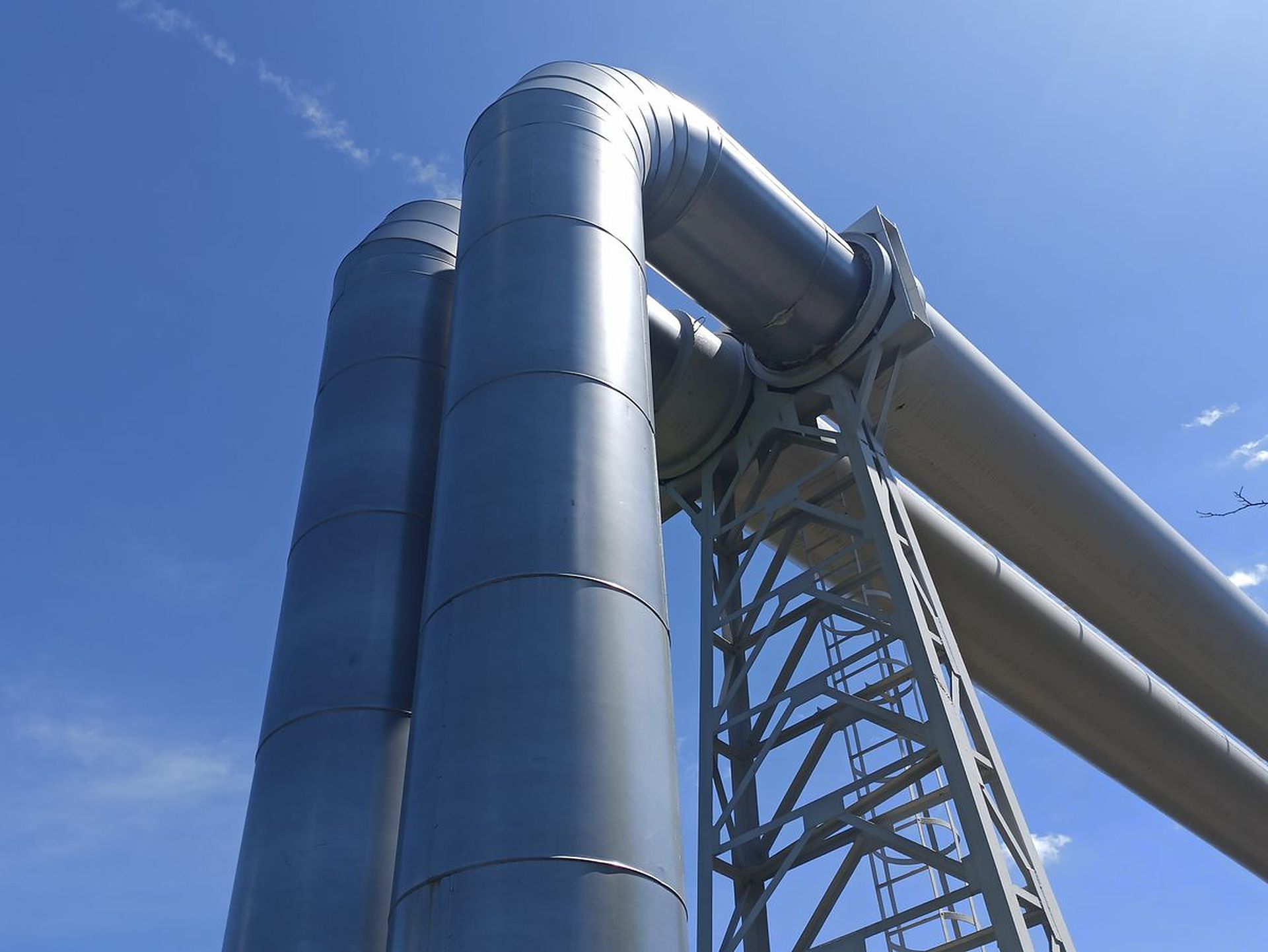 Large insulated steel pipes arching over a support tower against a blue sky